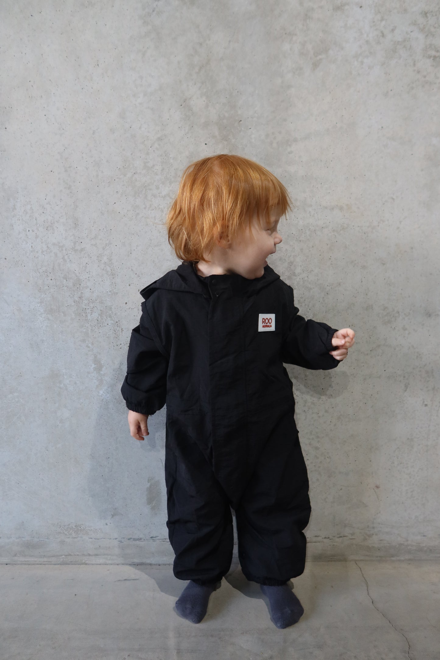 Toddler looking to the side in Roo Australia black windbreaker boiler suit, smiling against concrete wall.