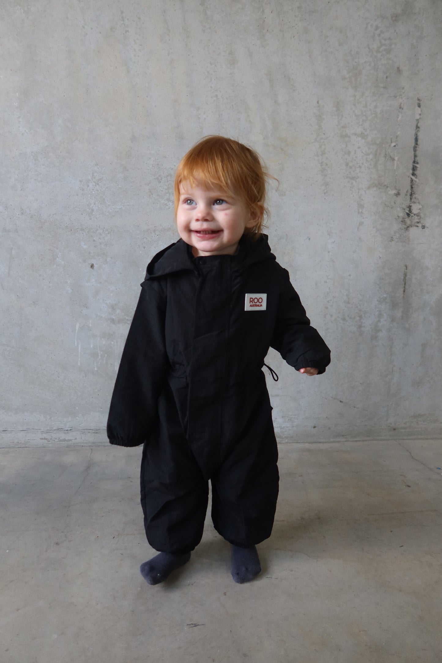 Child wearing Roo Australia’s black Windbreaker Boiler Suit, smiling while standing against a concrete wall.
