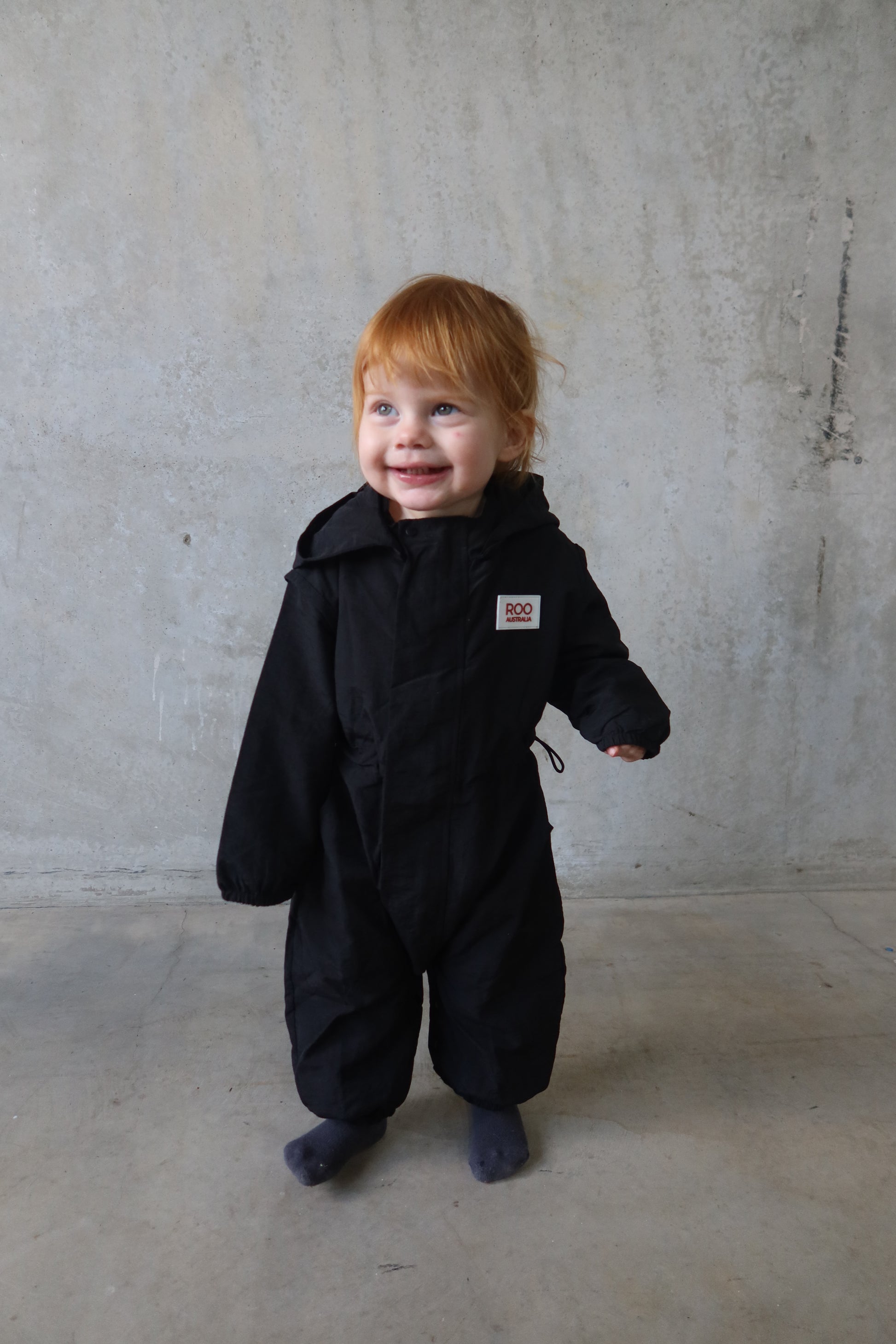 Child wearing Roo Australia’s black Windbreaker Boiler Suit, smiling while standing against a concrete wall.