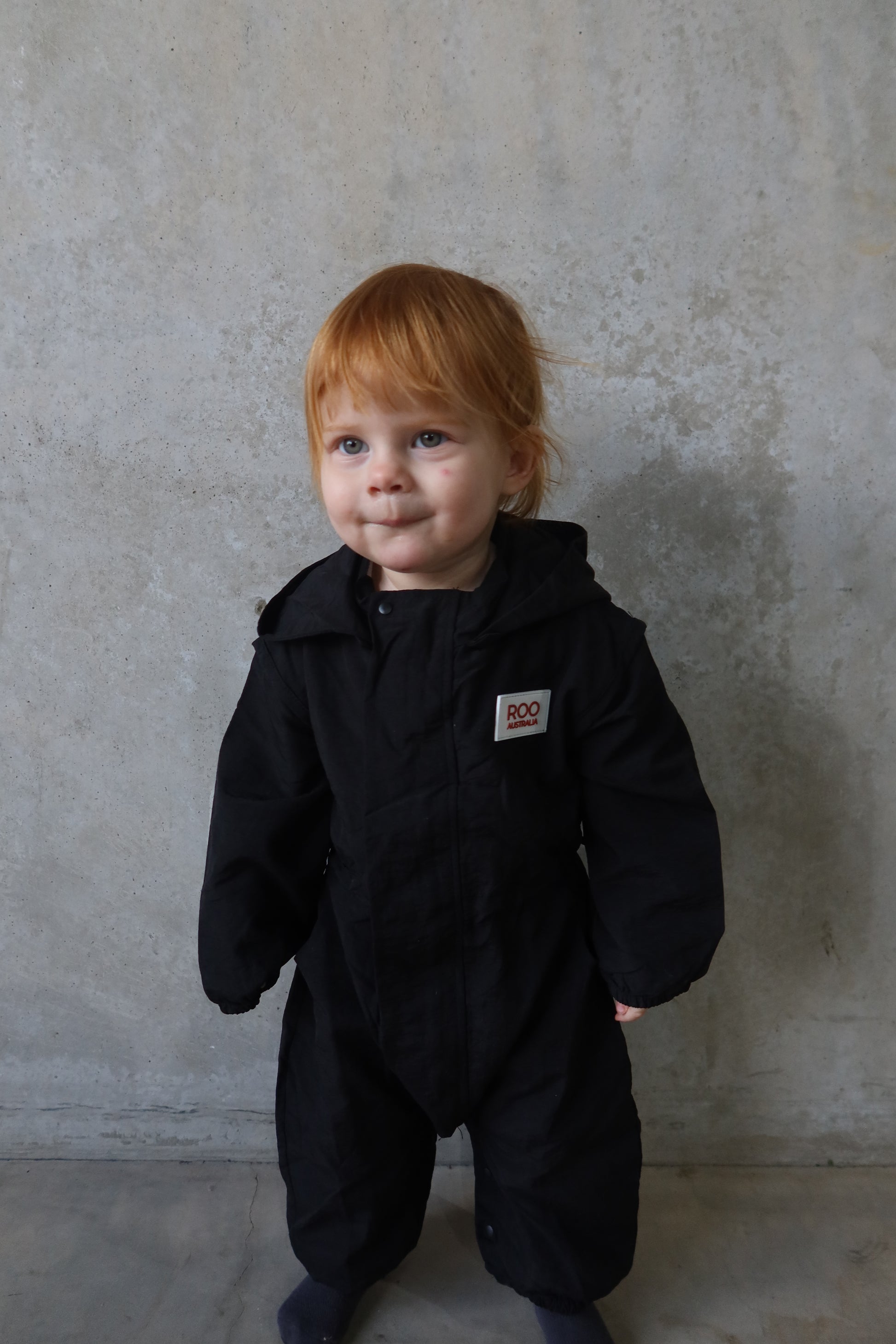 Child wearing black windbreaker boiler suit, standing and smiling.